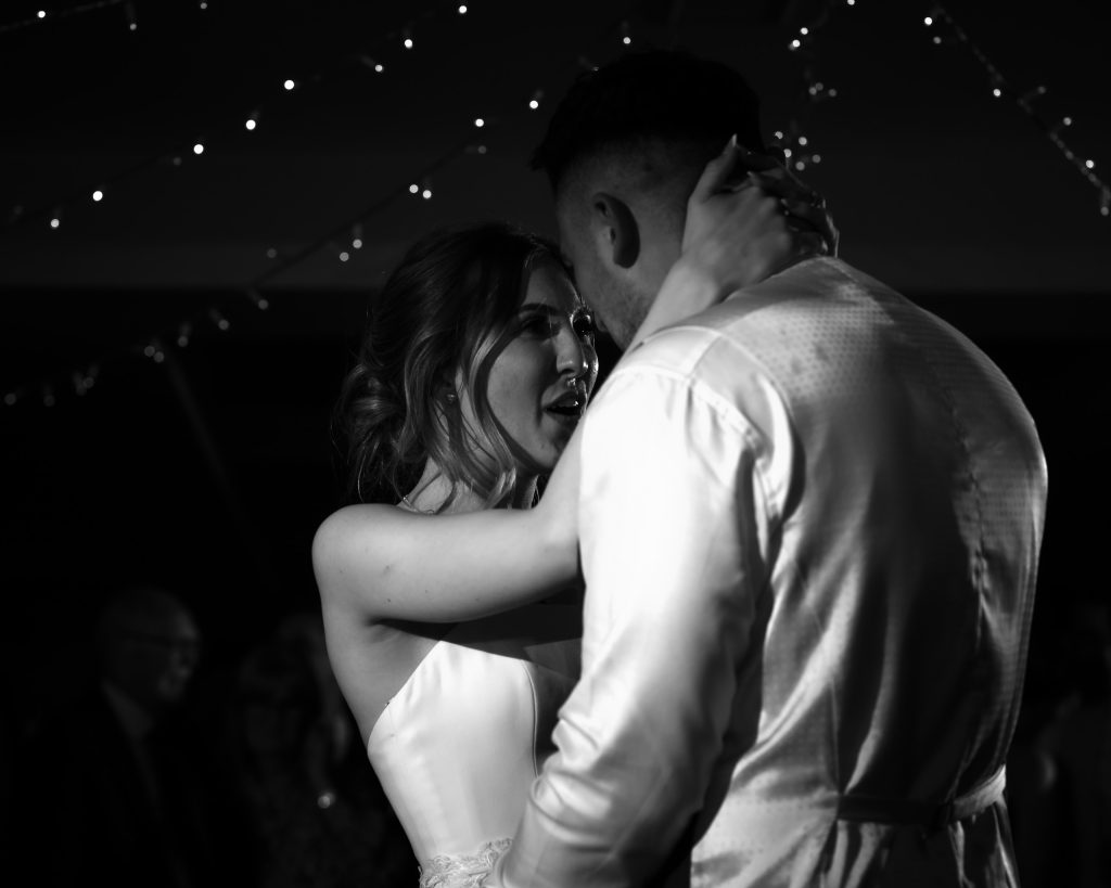 Black and white photo of a bride and groom holding each other close during their first dance under fairy lights