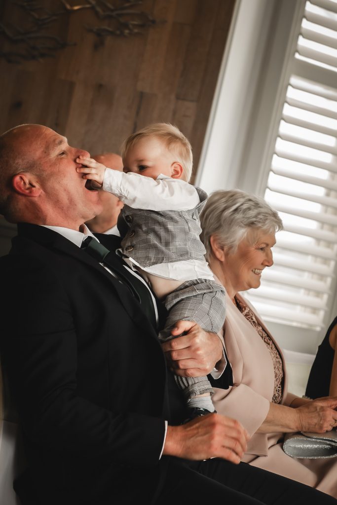 A toddler in a checked waistcoat puts his hand over a laughing guest’s mouth as the woman beside them smiles