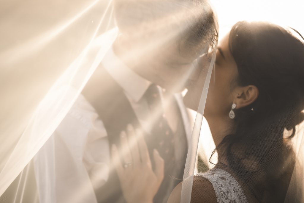 Bride and groom kissing beneath a sunlit veil