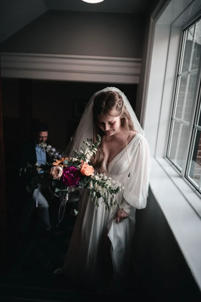 Bride smiling as she walks down the stairs with a colourful bouquet, window light beside her