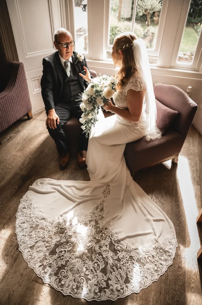 Bride and her father sitting together by a bright window before the ceremony, her lace train across the floor
