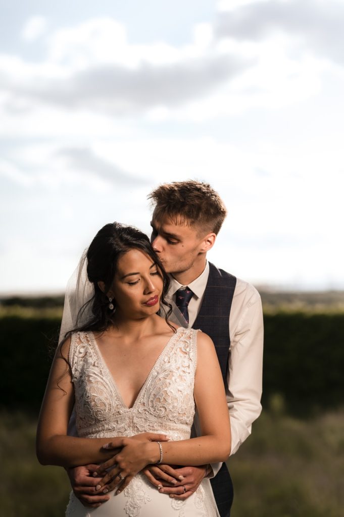Groom kissing the bride’s temple as she holds his hands against an open sky