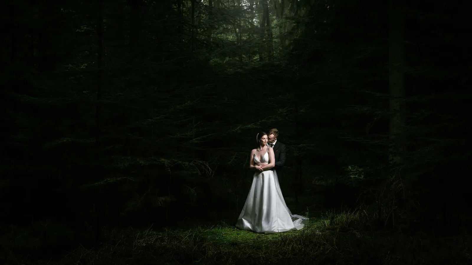 Bride and groom embracing in a pool of light in a dark pine forest