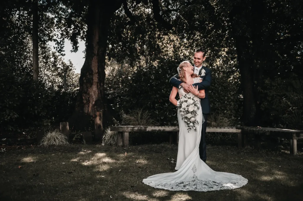 Groom embracing the bride from behind under tall trees, her lace train spread across the lawn