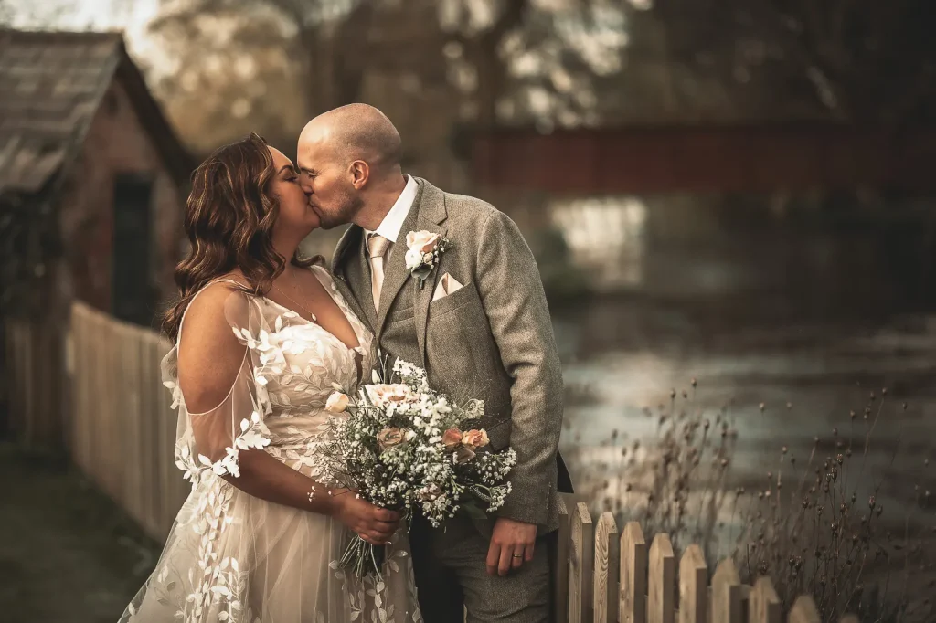 Bride and groom kissing beside a wooden fence and river, the bride holding a wildflower bouquet