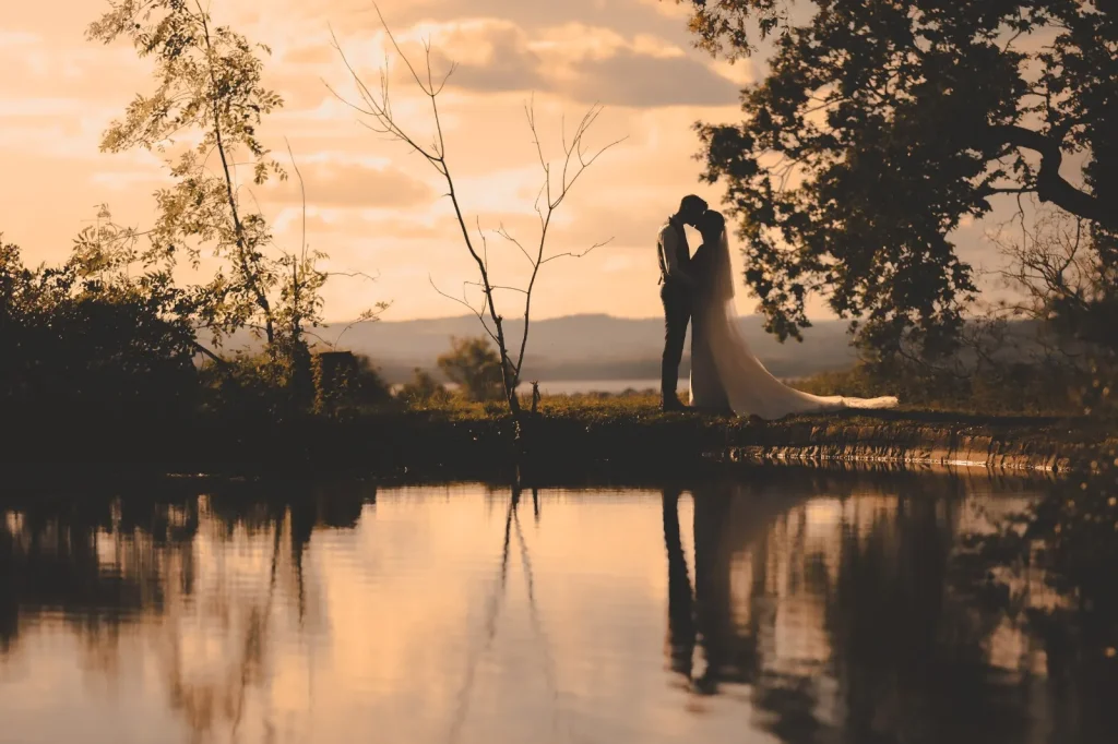 Bride and groom silhouetted at sunset beside a lake, reflected in the still water