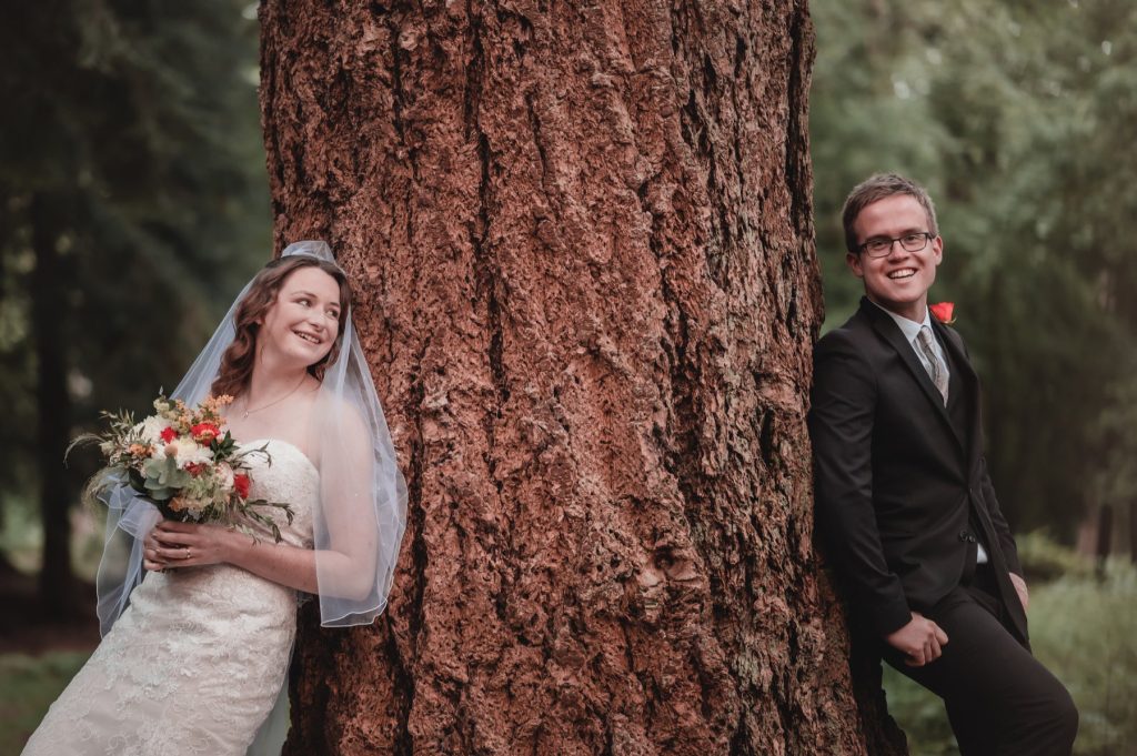 Bride laughing as she leans against a large pine while the groom leans on the other side of the trunk