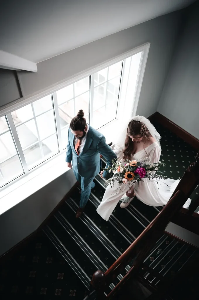 Bride and groom walking down a staircase beside a tall window, seen from above