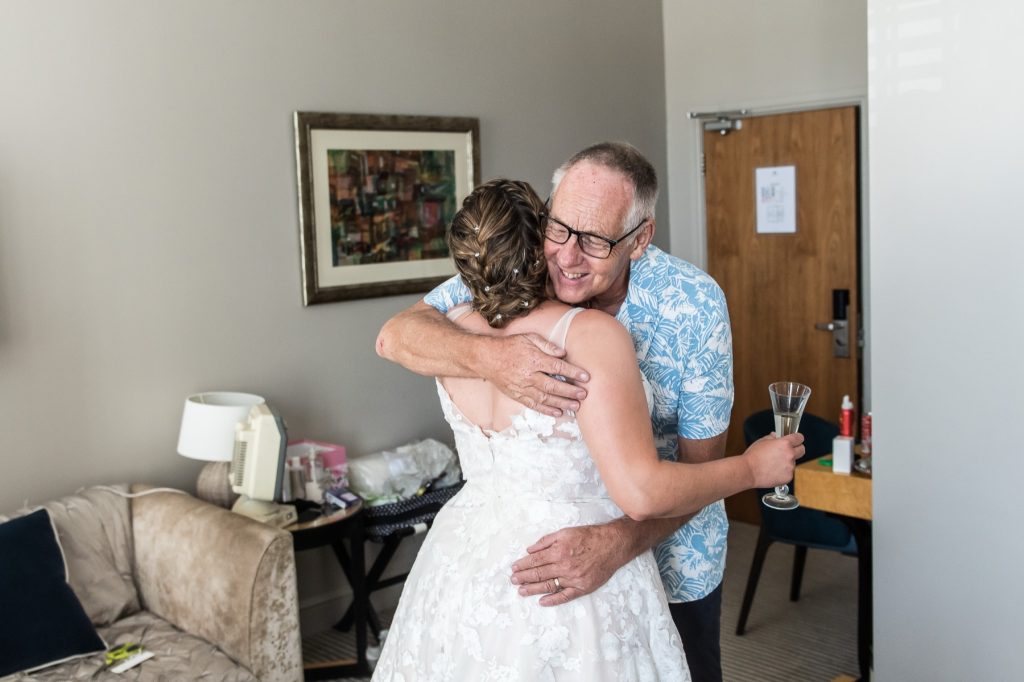 Bride hugging a guest in a blue floral shirt during morning preparations