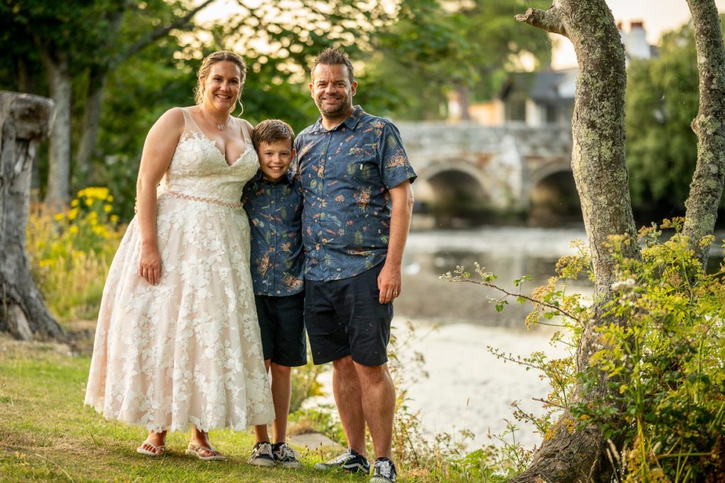 Bride, groom and a young boy standing by a river with a stone bridge behind them