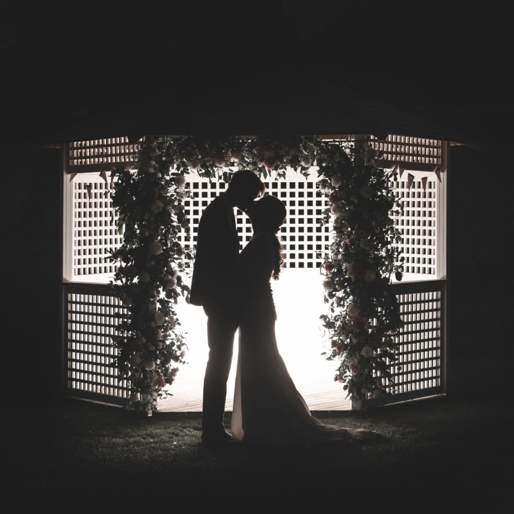 Bride and groom silhouetted in a kiss inside a white lattice gazebo