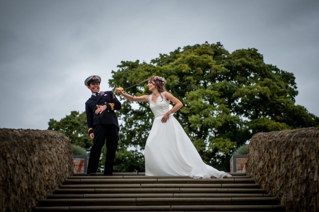 Groom in dress uniform and bride holding hands at the top of stone steps beneath a large tree