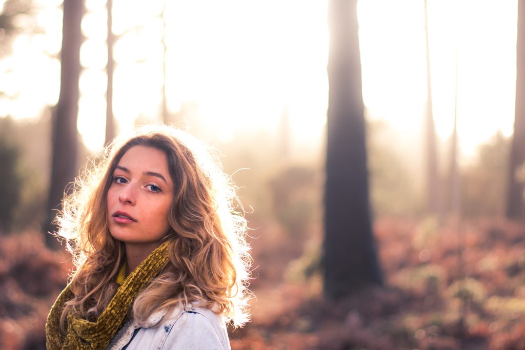 Young woman with long wavy hair and a mustard scarf, backlit by soft spring sunshine in a misty pine woodland