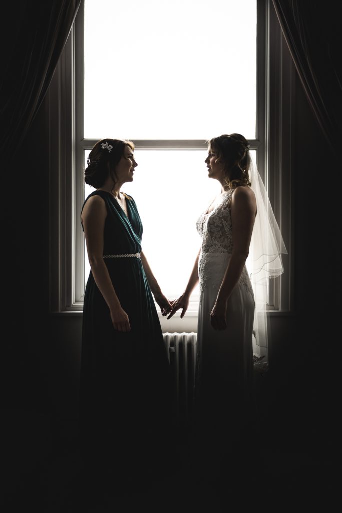Two brides holding hands, silhouetted against a tall window