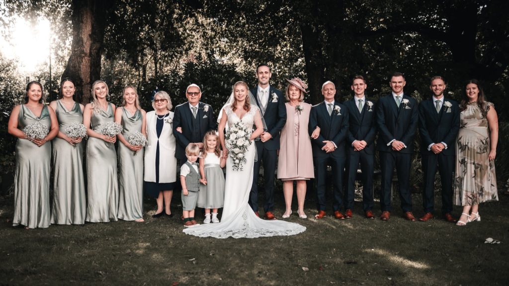 The full wedding party lined up on a lawn in front of tall trees
